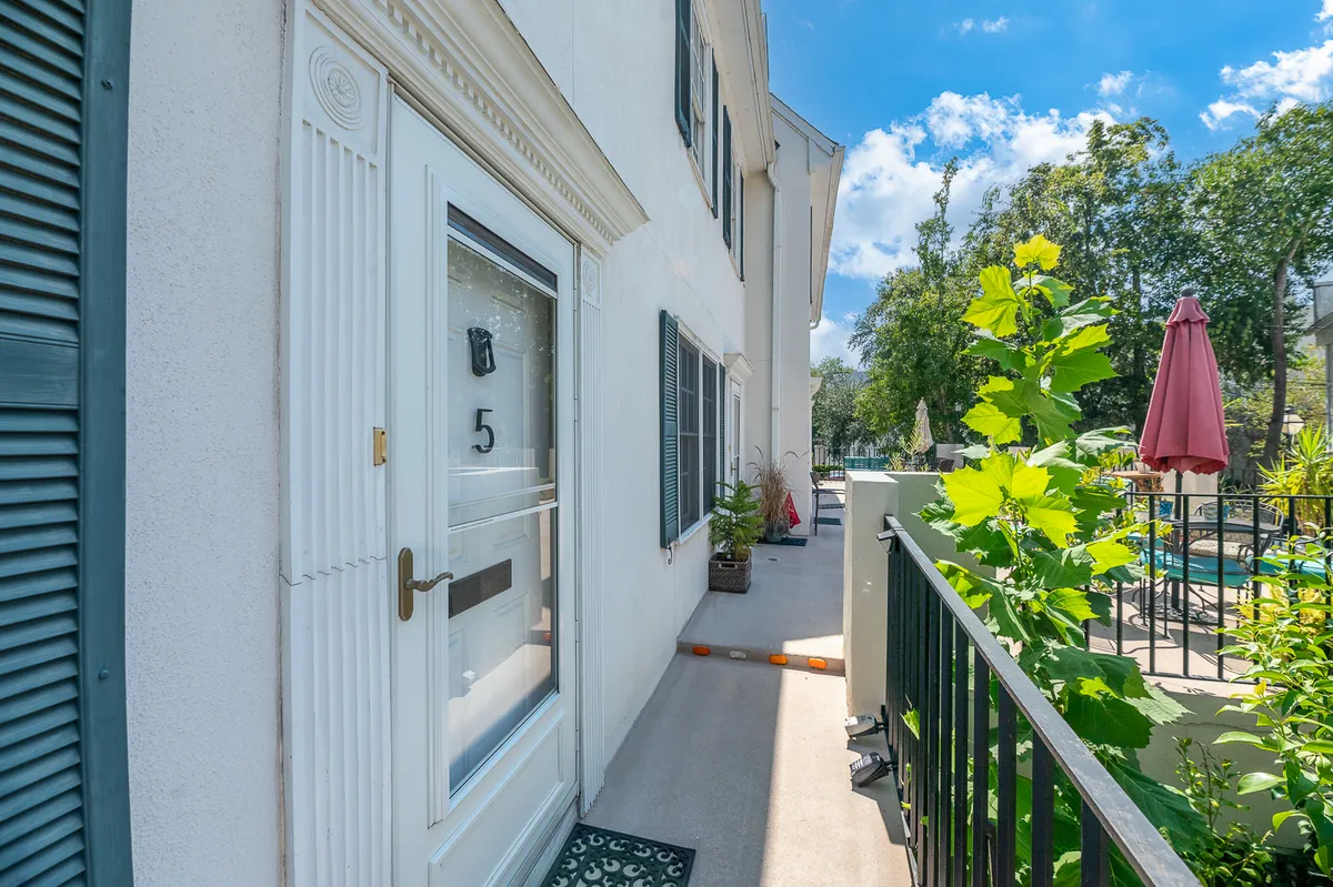 This image shows the inviting entrance to the property, framed by lush green foliage, with a front door leading to a private patio area. In the background, a sparkling pool and a patio umbrella suggest options for outdoor dining and relaxation. The scene is bright, welcoming, and hints at a seamless indoor-outdoor lifestyle.