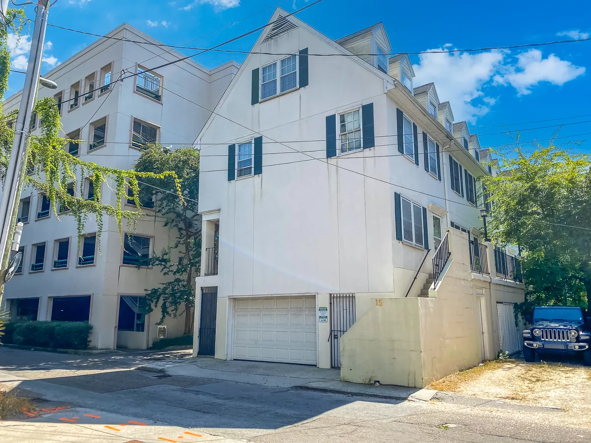 This exterior shot captures the white townhouse with green shutters, garage, and surrounding vibrant greenery. The perspective shows the property as nestled in a tranquil, walkable neighborhood—an inviting base for exploring nearby Charleston hotspots.