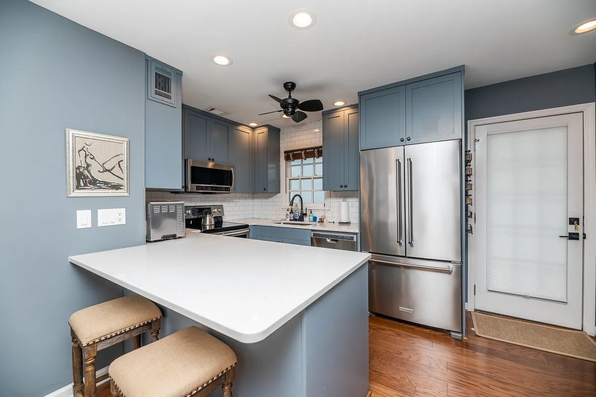 A modern kitchen and dining area with a central island, stainless steel appliances, blue cabinetry, and a sunlit window. The setup encourages gatherings over meals, casual dining, or even working at the island, all illuminated by ample natural light.