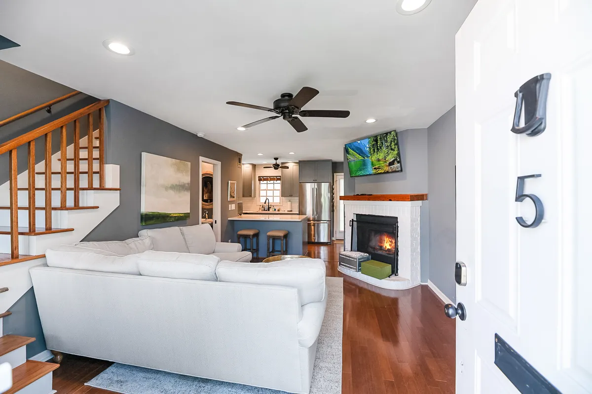 The image showcases a warm and inviting open-concept living area featuring a plush sofa arranged around a cozy fireplace, a modern kitchen island, and a stylish staircase leading upstairs. The space is bright yet intimate, perfect for relaxing, entertaining, or spending quality time with friends and family. The television and comfortable seating set the mood for movie nights after a day of downtown Charleston adventures.